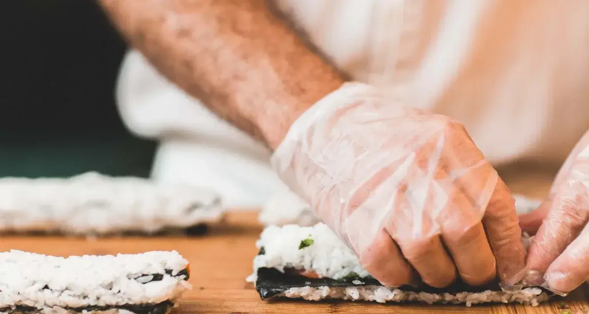 Ręce w rękawiczkach zwijają sushi na desce. Dowiedz się, jak zrobić zaprawę do ryżu sushi, by uzyskać idealny smak.