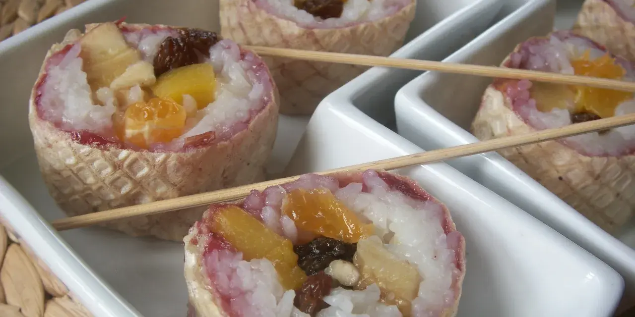 Słodkie sushi owocowe w wafelkach, z ryżem i kawałkami owoców, podane na białych tacach.