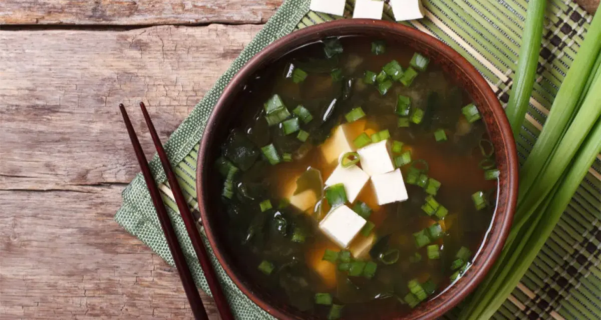Aksamitne miso z tofu, wodorostami i szczypiorkiem, podane z pałeczkami na drewnianym blacie.