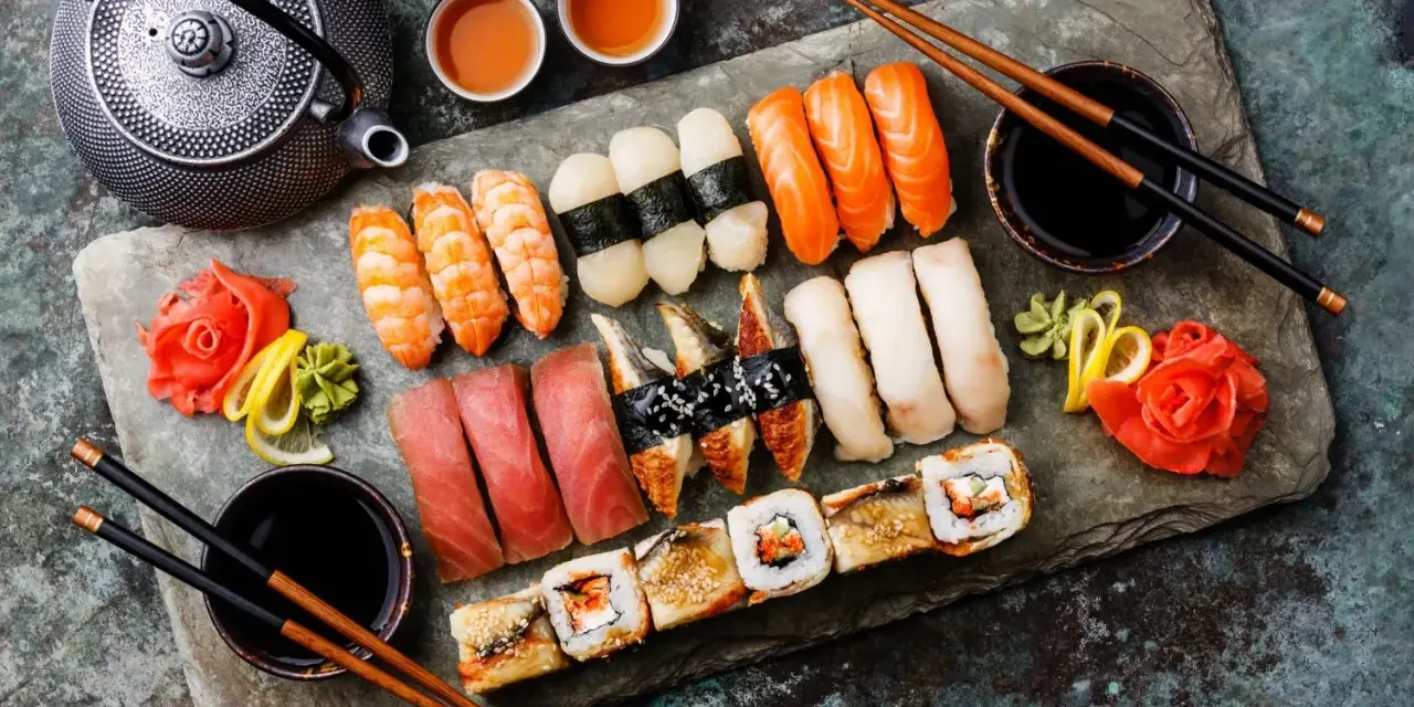Piękne składniki na sushi: nigiri z łososiem, krewetkami, tuńczykiem, węgorzem i rybą maślaną, a także rolki. Obok czajniczek, sos sojowy i marynowany imbir.