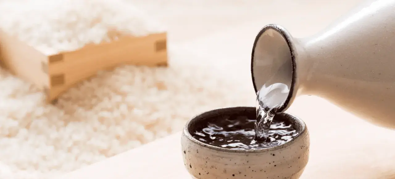Sake przelewana z dzbanka do czarki, w tle ryż. Poznaj, jak smakuje sake.