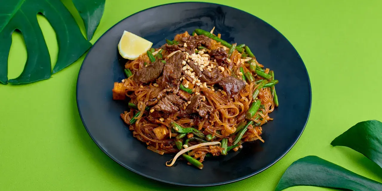 Pyszne pad thai z wołowiną, fasolką szparagową i orzeszkami na czarnym talerzu, ozdobione ćwiartką cytryny i liśćmi monstery.