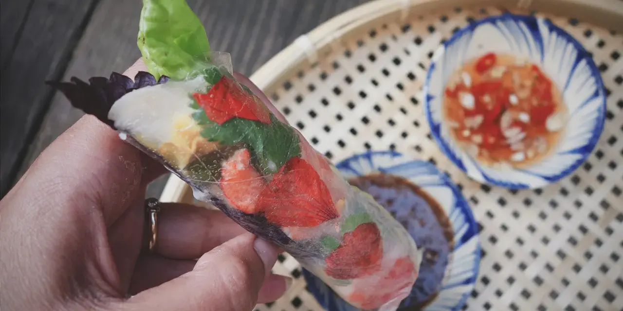 Ręka trzyma świeże rolls z papieru ryżowego z truskawkami i ziołami. Obok miseczki z sosem.