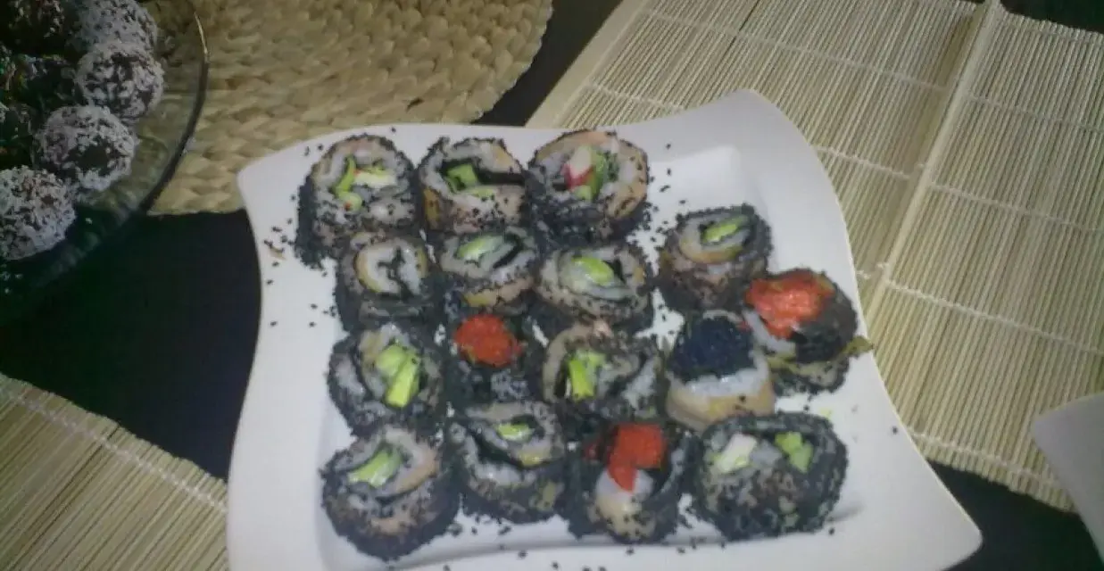 Talerz pełen sushi z sezamem, z widocznymi kawałkami awokado i ikrą. Obok deserki w wiórkach kokosowych.