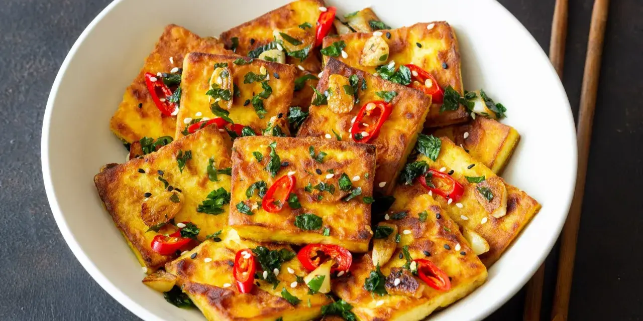 Chrupiące tofu w tempurze, posypane sezamem, chili i natką pietruszki, podane w białej misce z pałeczkami.
