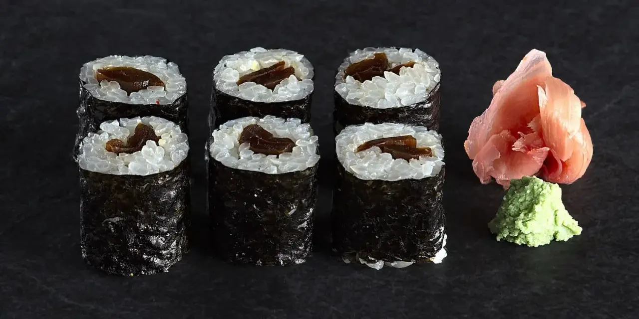 Sześć roladek sushi z ryżem i ciemnym nadzieniem, prawdopodobnie sushi z tykwą. Obok różowy imbir i zielone wasabi.