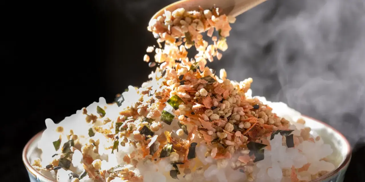 Łyżka sypie na parujący ryż kolorowy furikake. To japońska posypka, która dodaje smaku.
