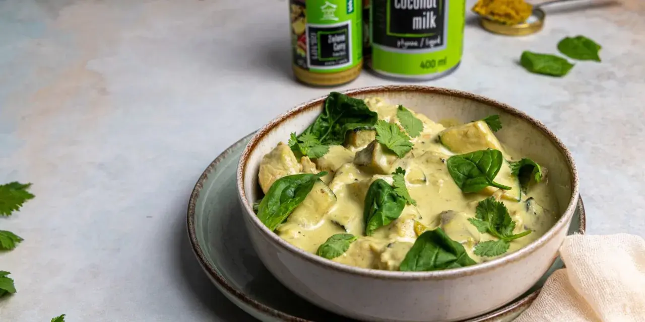 Aksamitny kurczak w zielonym curry, udekorowany świeżym szpinakiem i kolendrą. Obok puszka mleka kokosowego i słoiczek pasty curry.