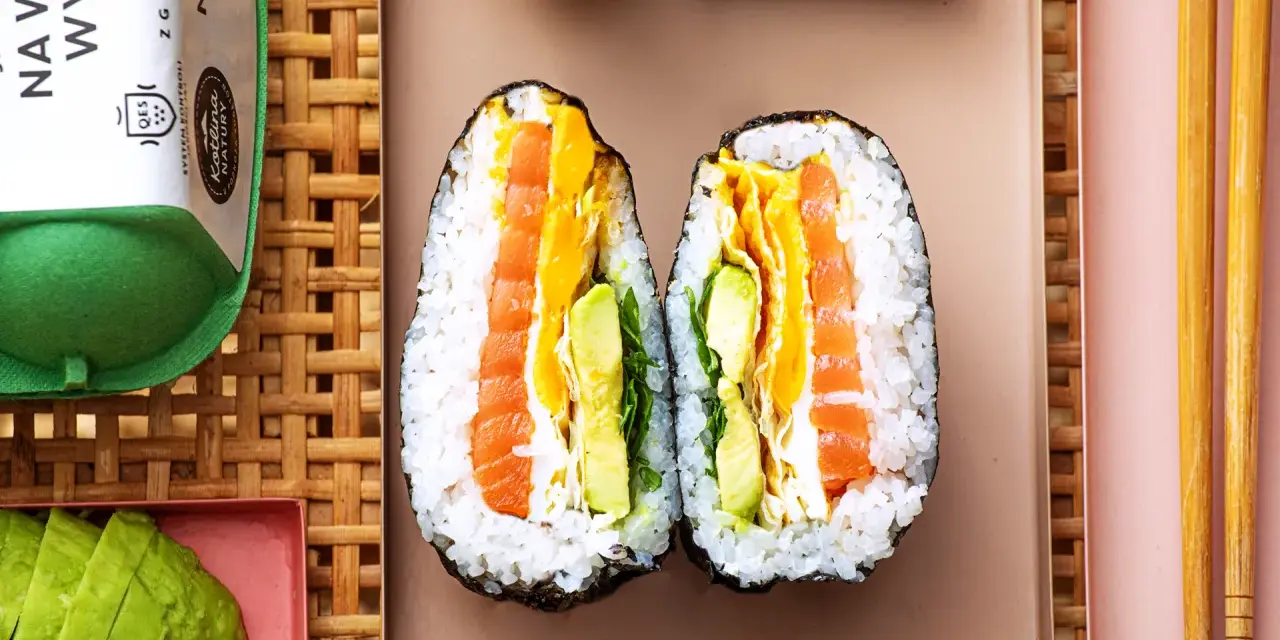 Dwa połówki onigiri z łososiem, awokado i jajkiem, podane na talerzu z pałeczkami.
