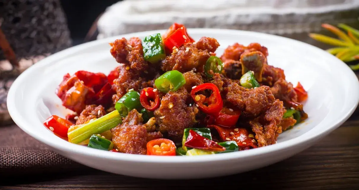Chrupiący kurczak po kantonsku z papryczkami chili i zieloną cebulką na białym talerzu.