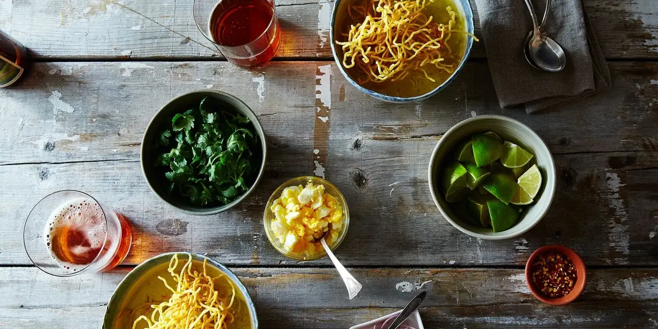 Dwa miseczki khao soi, świeża kolendra, limonki, jajko na twardo, czerwona cebula i płatki chili na drewnianym stole.