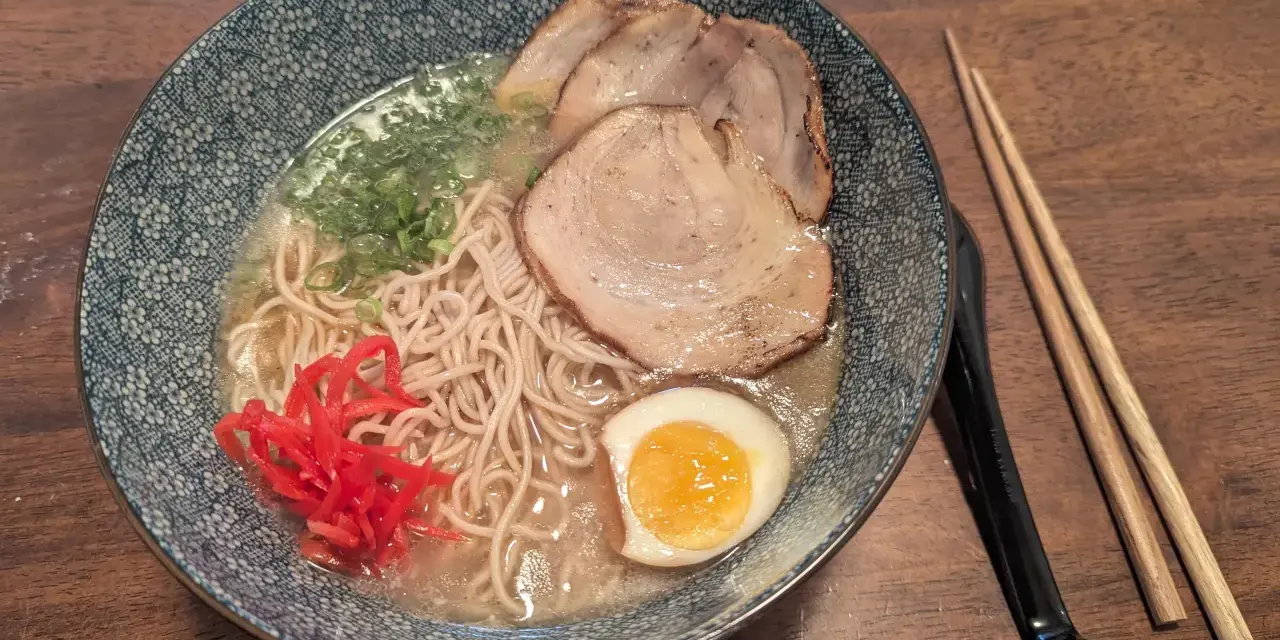 Miseczka z ramen: makaron, plastry mięsa, jajko na pół, zielona cebulka i czerwone przyprawy do ramen. Obok pałeczki.
