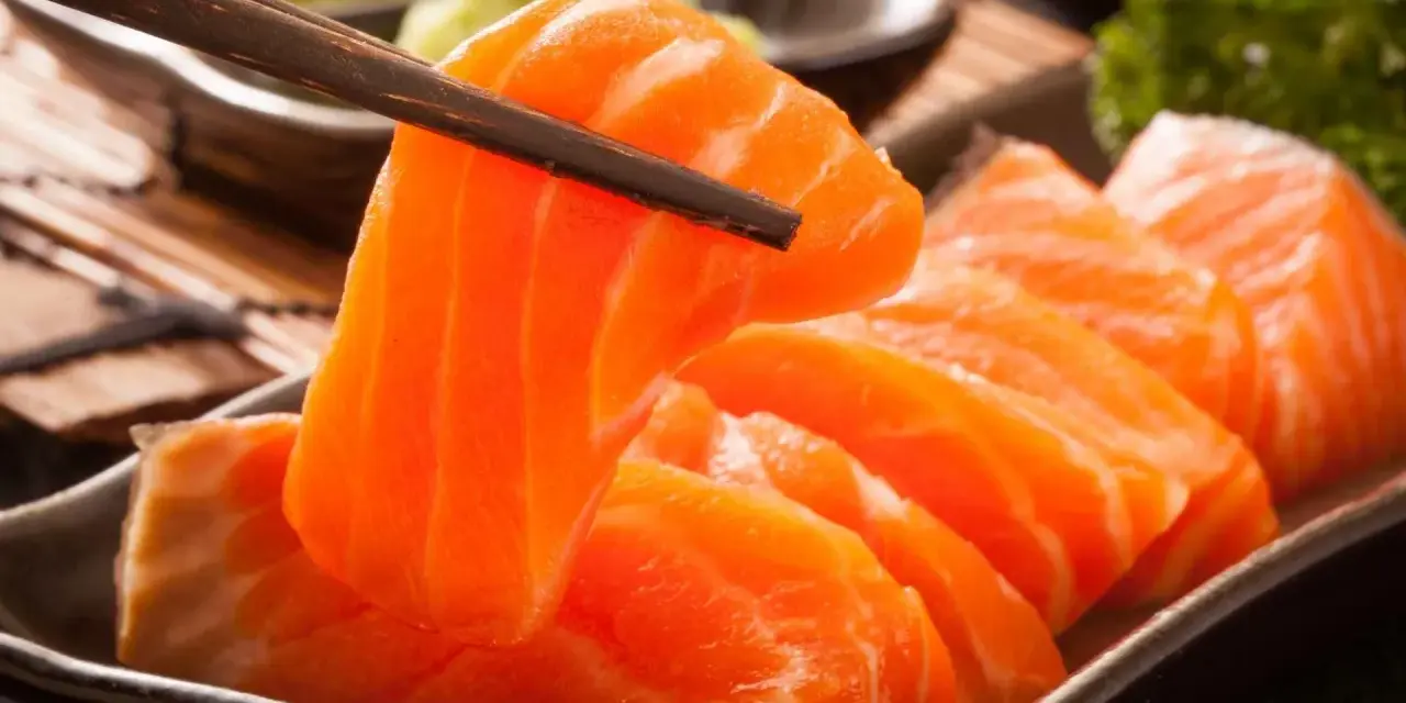 Sashimi co to? Świeże, pomarańczowe kawałki łososia podniesione pałeczkami, obok zielonej pasty wasabi i natki pietruszki.