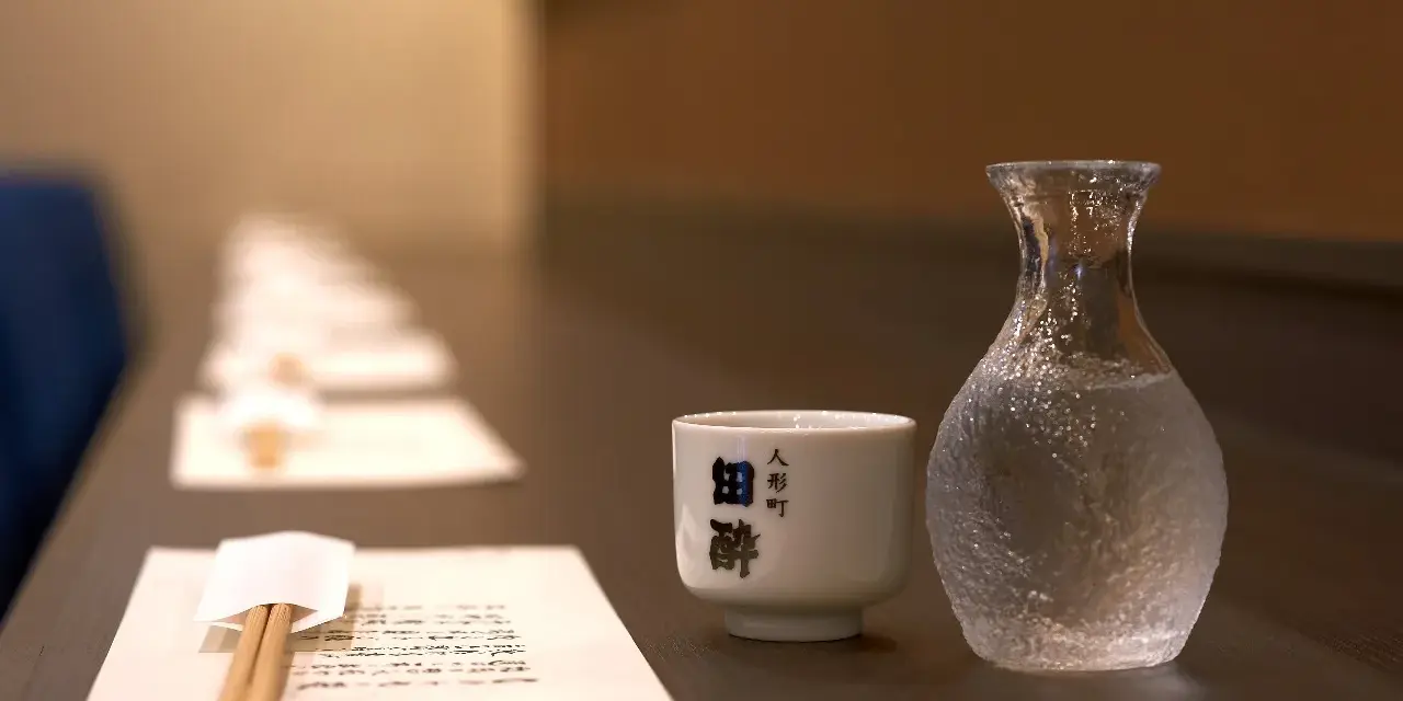 Sake ile procent? Delikatna czarka i karafka z sake czekają na degustację. W tle rząd nakryć.