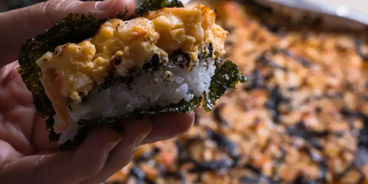Dłoń trzyma kawałek sushi bake z ryżem, nori i zapiekanym farszem.