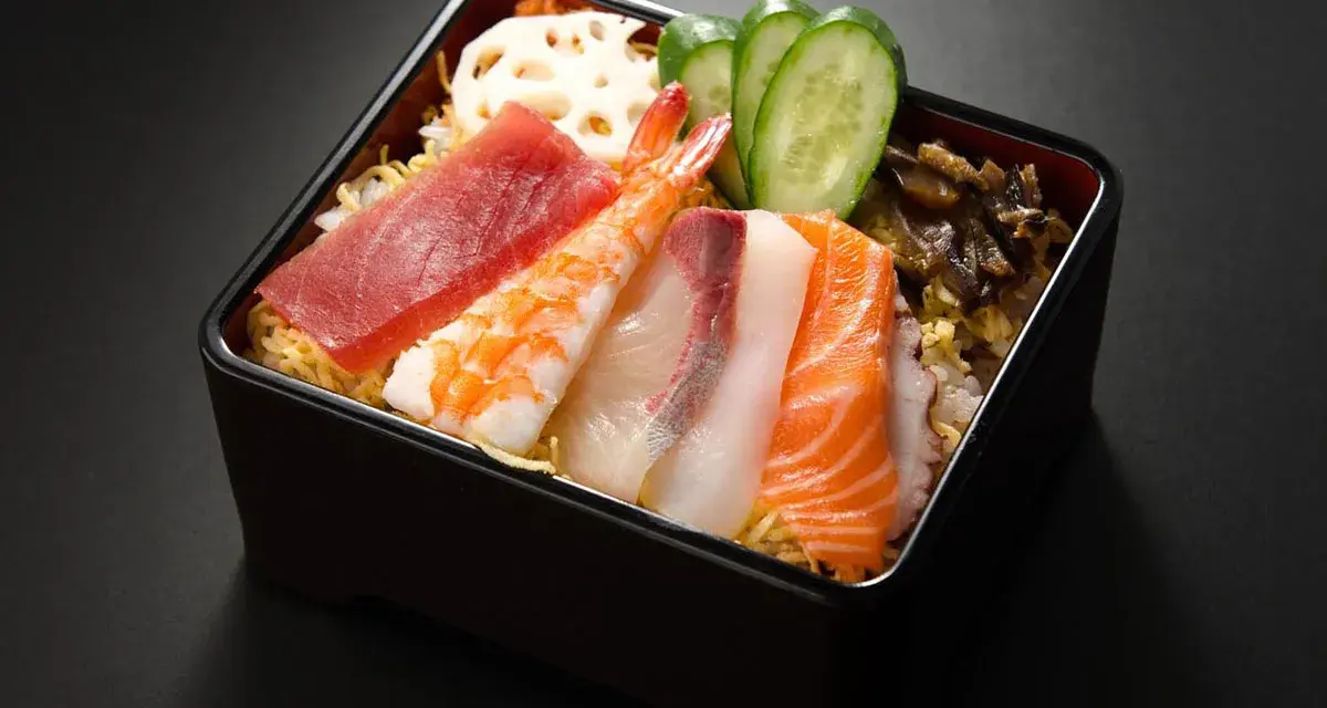 Chirashi zushi: kolorowe plastry ryb (tuńczyk, łosoś, biała ryba), krewetka, plasterki ogórka i lotosu na ryżu.