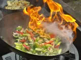 Warzywa w woku płoną w ogniu, tworząc aromatyczny stir fry. W tle widać drugie danie.