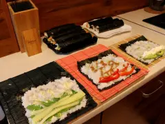 Przygotowanie ryżu do sushi. Na matach leżą arkusze nori z ryżem i dodatkami: ogórkiem, awokado, papryką i sosem.