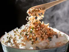 Łyżka sypie na parujący ryż kolorowy furikake. To japońska posypka, która dodaje smaku.
