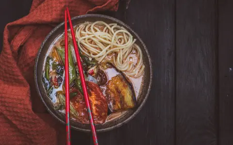 A miseczce z aromatycznym ramen wegetariańskim, pełnej makaronu, grillowanego bakłażana i pomidorów, spoczywają czerwone pałeczki.