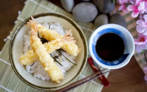 Chrupiące krewetki w cieście tempura na ryżu, obok miseczka z ciemnym sosem do tempury.