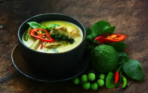 Aromaticzne zielone curry z kawałkami mięsa, papryczką chili, zielonym pieprzem i liśćmi bazylii. Obok miski świeże składniki: limonka kaffir, pomidorki i papryczki chili.