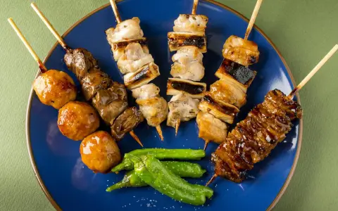 Różnorodne szaszłyki kurczak yakitori: mięso, wątróbka, kulki mięsne i warzywa na niebieskim talerzu.