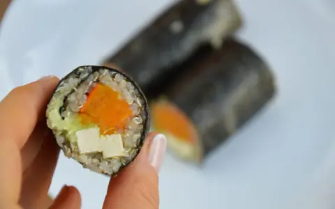 Rolka sushi z tofu, dynią i awokado, trzymana w dłoni.