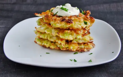 Stos placków okonomiyaki, polanych śmietaną i posypanych natką pietruszki, na białym talerzu.