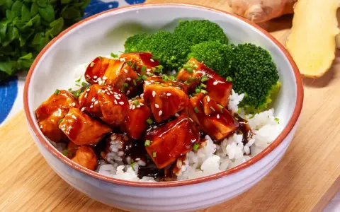 Miseczka ryżu z kawałkami lśniącego lososia teriyaki, posypanego sezamem i szczypiorkiem, obok parujących różyczek brokułów.