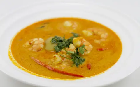 Aksamitna zupa tom yum z krewetkami, świeżą kolendrą i kawałkami chili na białym talerzu.