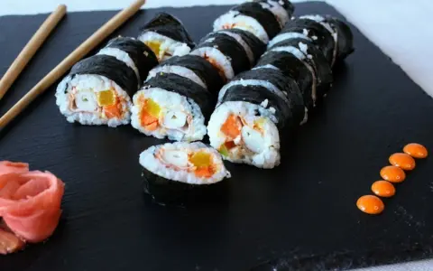 Roladki sushi z surimi, ryżem i warzywami na czarnej desce, obok pałeczki i różowy imbir.