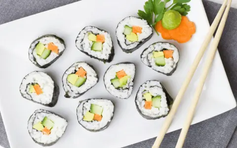 Talerz z rolkami sushi z awokado, ogórkiem i marchewką. Obok pałeczki i dekoracja z marchewki z wasabi. Idealna ilość octu do sushi podkreśla smak.