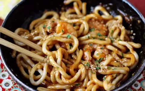 Pyszne udon z krewetkami w sosie, posypane sezamem i natką pietruszki. Danie w czarnej misce, gotowe do jedzenia pałeczkami.
