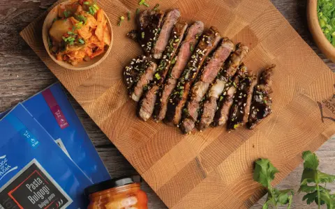 Pokrojony stek bulgogi polany sosem, obok miseczka z kimchi i opakowanie pasty do bulgogi.