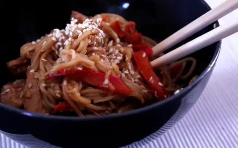 Miseczka pełna aromatycznego makaronu teriyaki z kawałkami kurczaka i papryką, posypana sezamem.