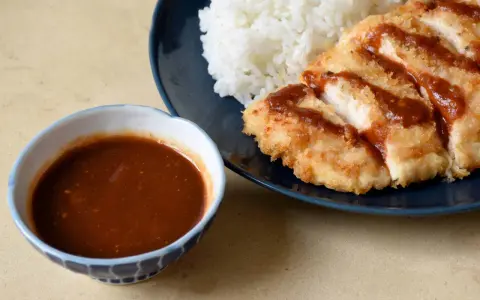 Chrupiący kotlet schabowy polany sosem tonkatsu, obok biały ryż i miseczka sosu.