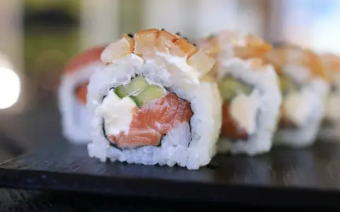 Uramaki co to? Rolka sushi z łososiem, awokado i krewetką, ryż na zewnątrz, na czarnej desce.