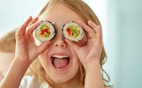 Uśmiechnięta dziewczynka trzyma kawałki sushi dla dzieci przed oczami, udając, że ma rolki zamiast oczu.
