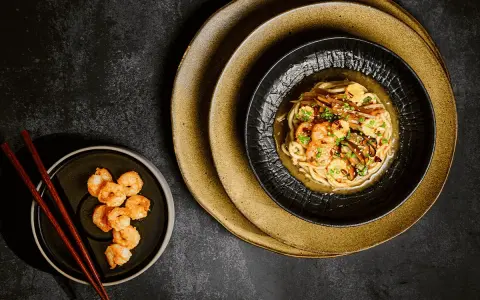 Chintan ramen z krewetkami i warzywami, podany na czarnym talerzu, obok miseczki z dodatkowymi krewetkami i pałeczkami.