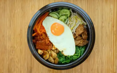 Apetyczne koreańskie jedzenie: miska bibimbap z jajkiem sadzonym, warzywami i mięsem na drewnianym tle.
