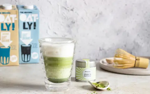 Warstwowy napój matcha z mlekiem kokosowym w szklance z podwójnymi ściankami. Obok puszka z matchą i bambusowa miotełka.