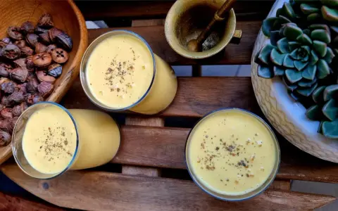 Trzy szklanki kremowego mango lassi, posypane przyprawami, obok miski z suszonymi owocami i sukulentu.