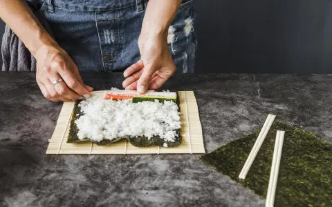 Ręce układają składniki do zwijania sushi na macie bambusowej. Ryż, paluszki krabowe i ogórek czekają na zawinięcie.