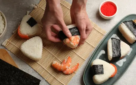 Ręce formują onigiri z krewetkami, tworząc pyszne japońskie jedzenie. Obok miska ryżu, nori i talerz gotowych przysmaków.