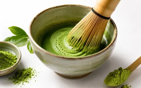 Tradycyjne matcha przygotowanie: bambusowa miotełka miesza zielony proszek z wodą w miseczce, tworząc piankę. Obok mała miseczka z proszkiem i drewniana łyżeczka.