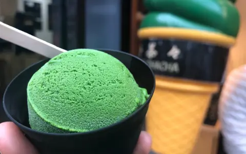 Zielone, japońskie lody matcha w czarnym kubeczku, z drewnianą łyżeczką. W tle wielki, żółty rożek z lodami.