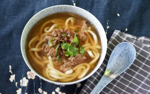 Gorąca zupa udon z wołowiną i szczypiorkiem, ozdobiona kolendrą, w misce na tle z motywem kwiatowym. Obok leży niebieska łyżka.
