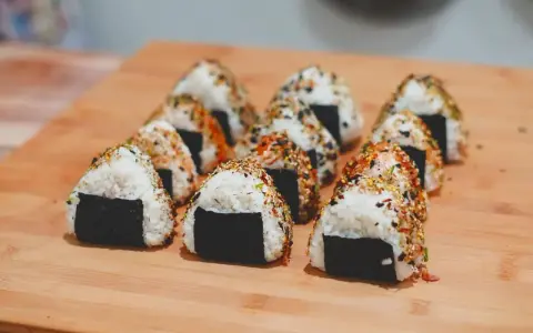 Rząd trójkątnych onigiri z kurczakiem, posypanych sezamem i przyprawami, ułożonych na drewnianej desce.