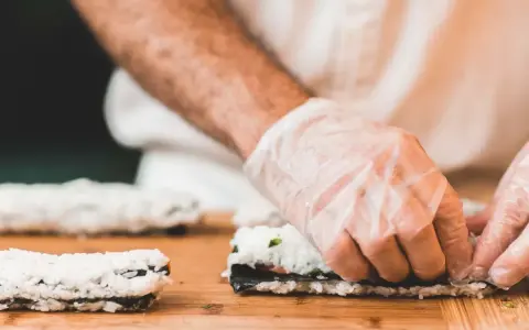 Ręce w rękawiczkach zwijają sushi na desce. Dowiedz się, jak zrobić zaprawę do ryżu sushi, by uzyskać idealny smak.